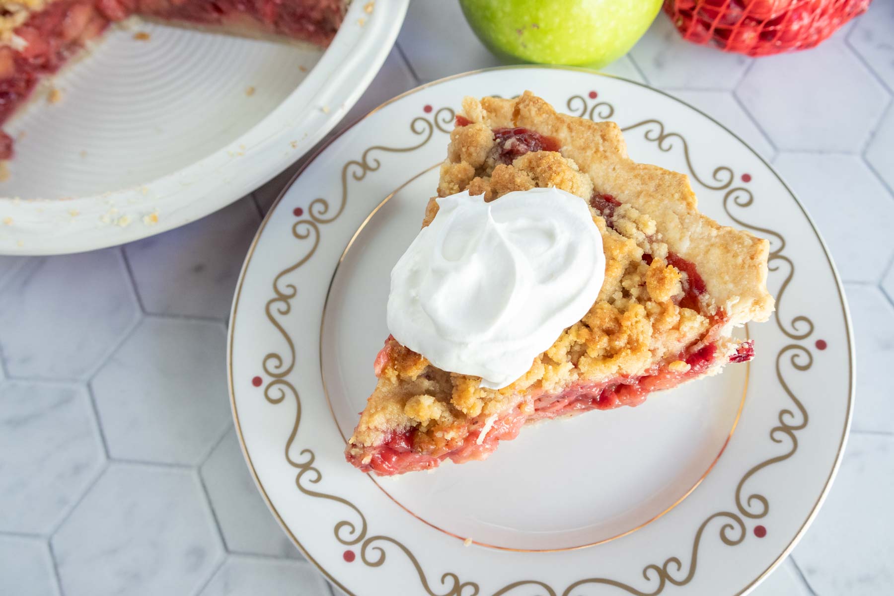 A slice of apple pie topped with whipped cream on a decorative plate, placed next to a whole pie and a green apple.