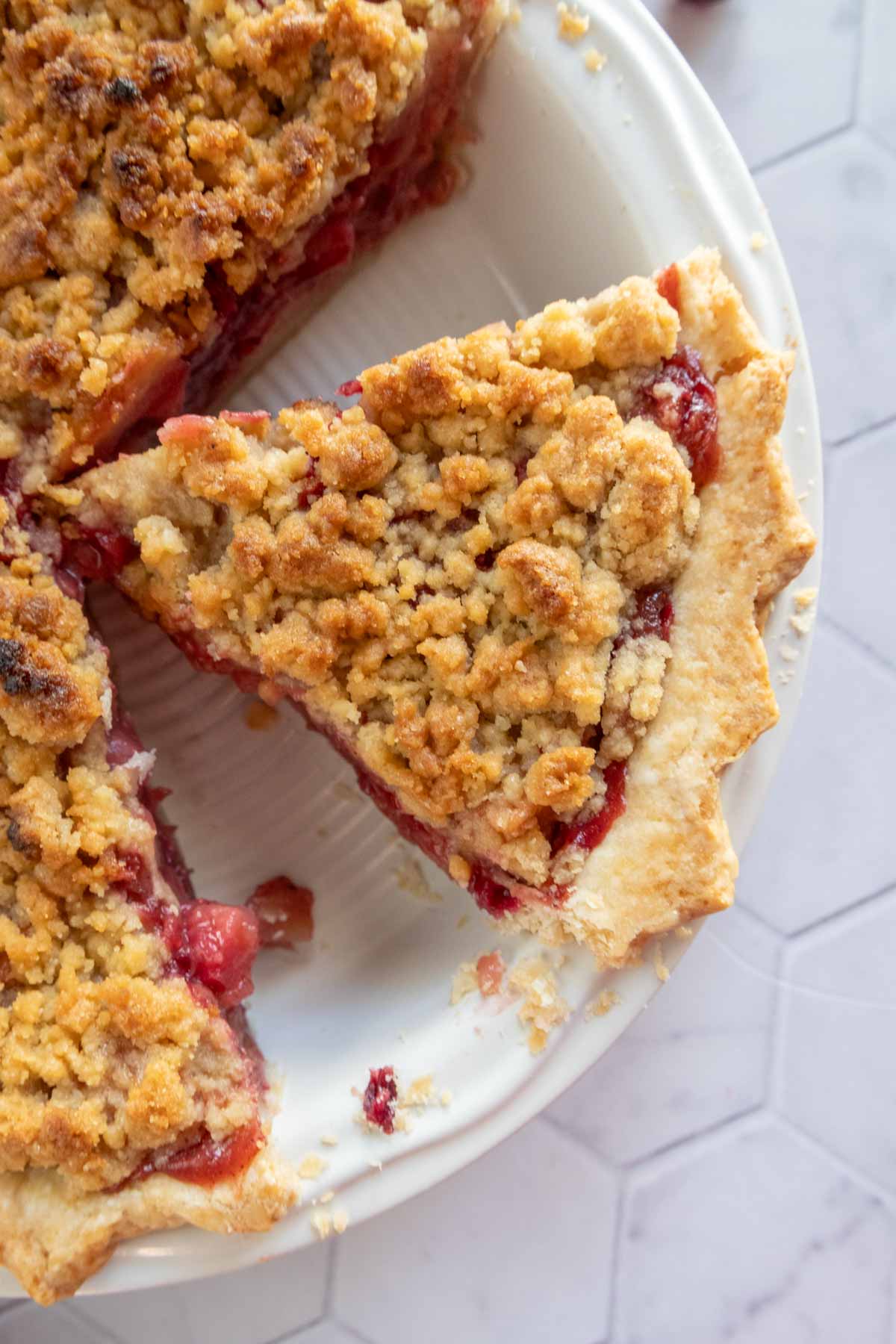 Slice of crumb-topped fruit pie on a white plate with hexagonal tile background.