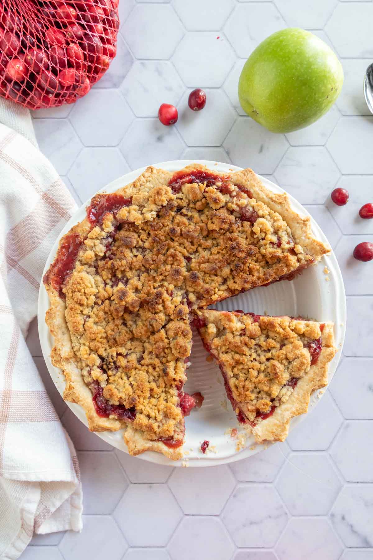 A baked pie with a crumb topping has one slice removed. An apple and cranberries are nearby on a hexagonal tile surface, next to a folded cloth.