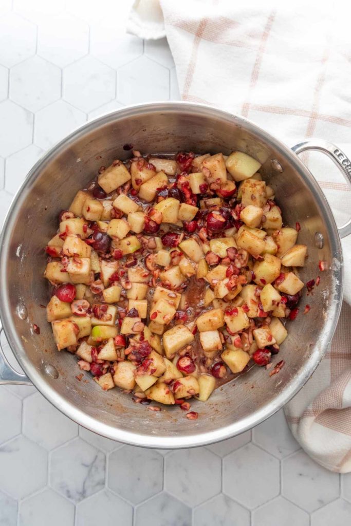 A stainless steel pot containing a mixture of diced apples and cranberries on a white hexagonal tile surface. A beige checked cloth is partially visible beside the pot.