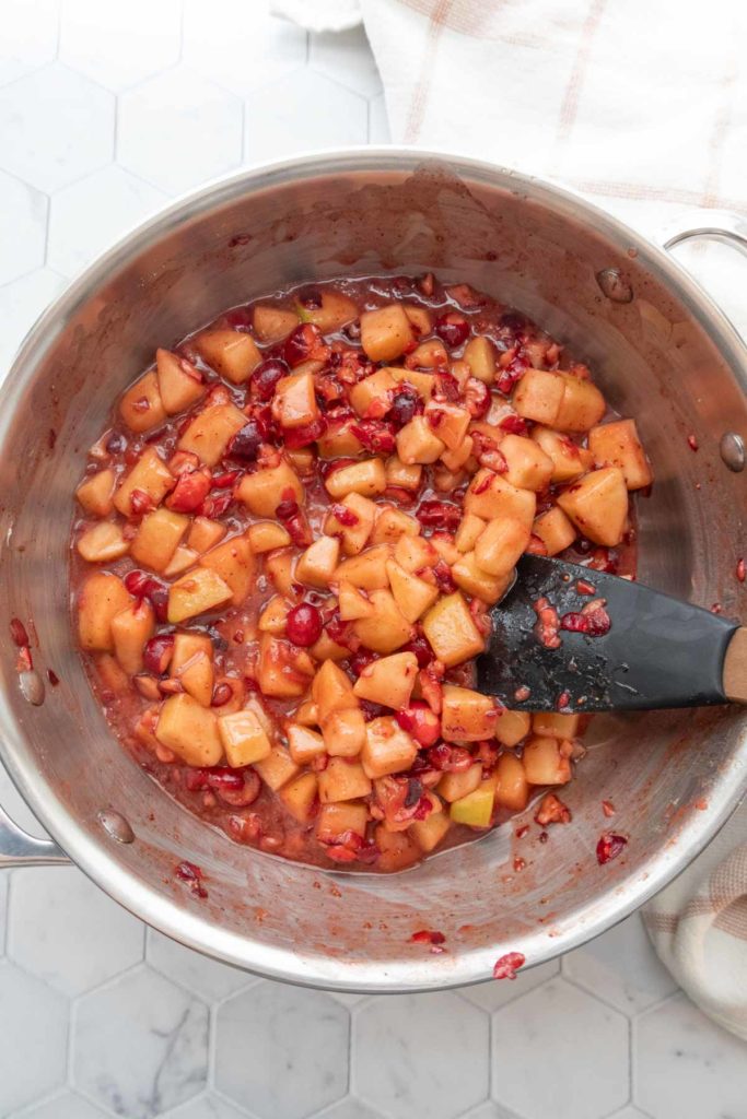 Chopped fruit, including apples and cranberries, being stirred in a pot with a black spatula on a white tiled surface.