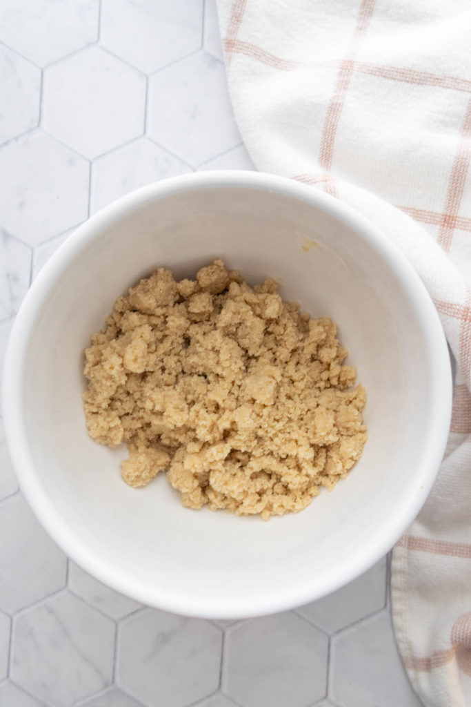 A white bowl on a hexagonal tiled surface contains crumbled cookie dough. A checked cloth is partially visible beside it.