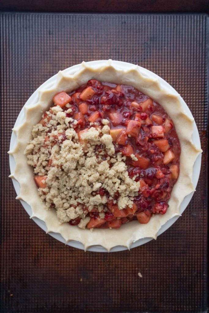 A partially assembled fruit pie with a crumb topping on one side and exposed fruit filling on the other, placed on a textured baking tray.