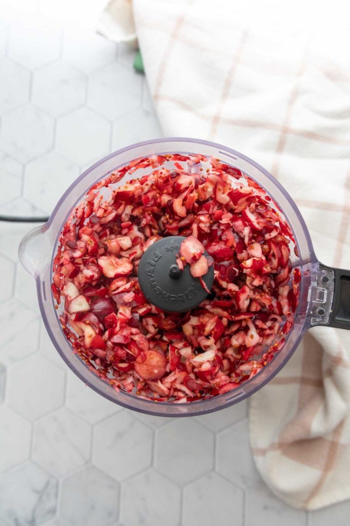 Chopped red ingredients in a food processor on a hexagonal tile surface next to a plaid fabric.