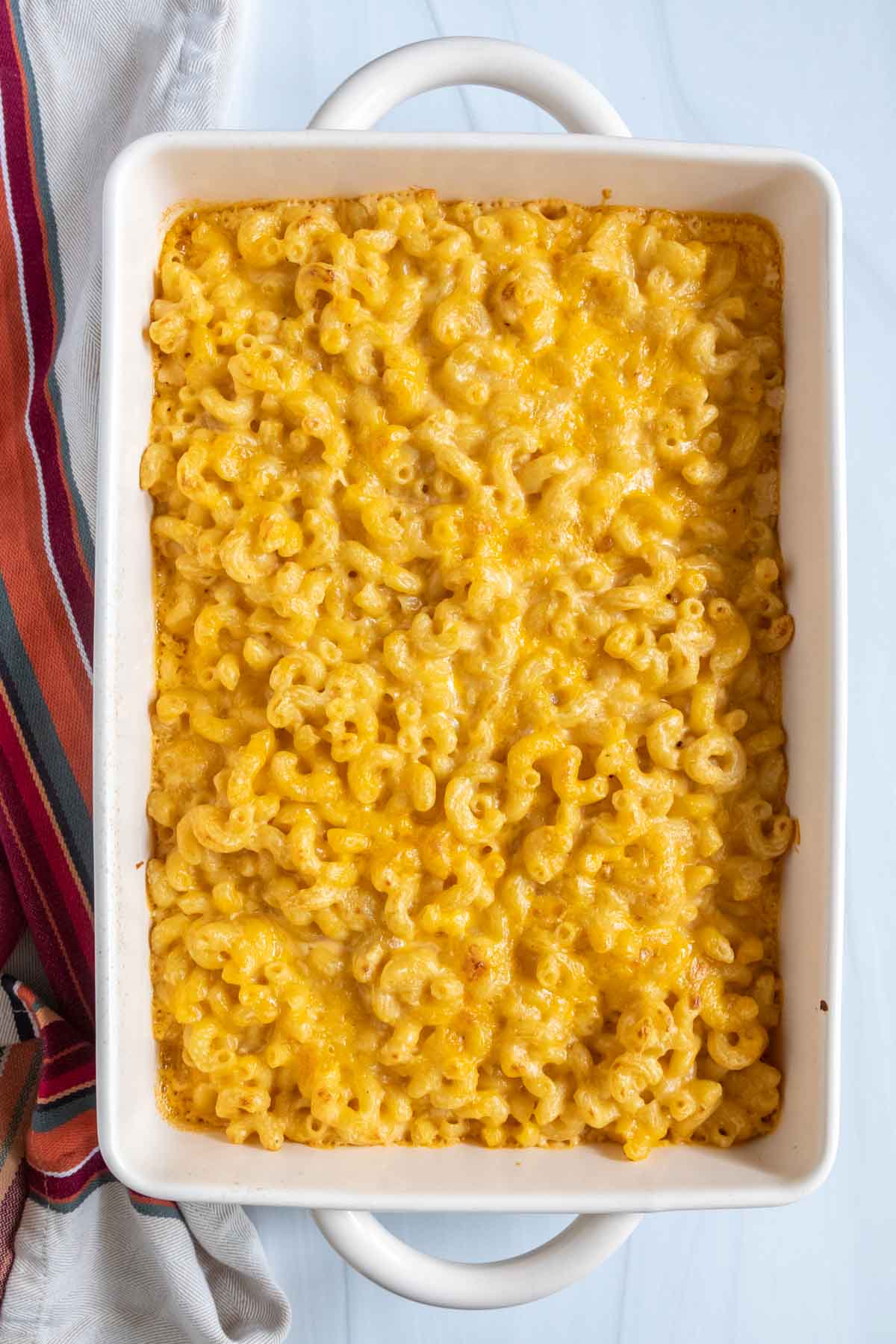 A baked dish of macaroni and cheese in a white rectangular baking dish, with a striped cloth beside it.