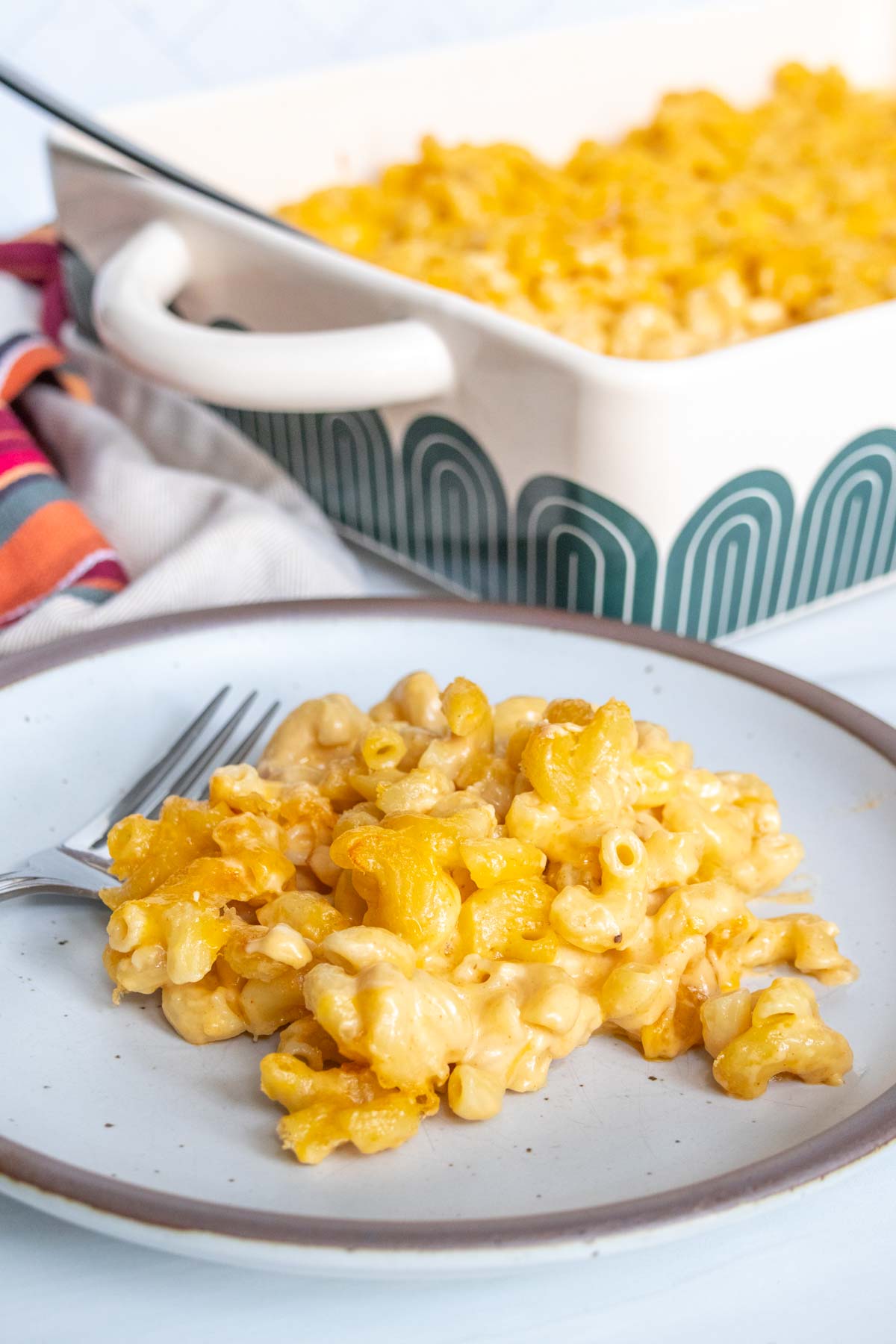 A plate with baked macaroni and cheese is in the foreground, and a casserole dish with more of the dish is in the background. A fork is placed beside the macaroni on the plate.