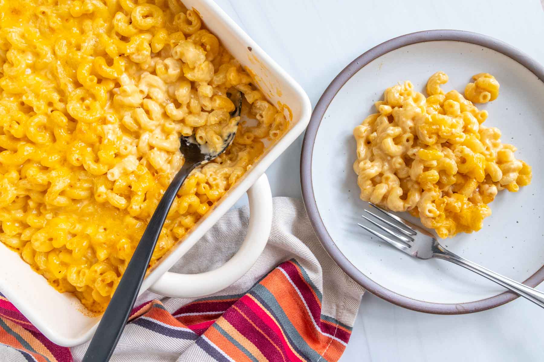 A serving of macaroni and cheese on a white plate next to a baking dish filled with more macaroni and cheese, with a striped cloth underneath.