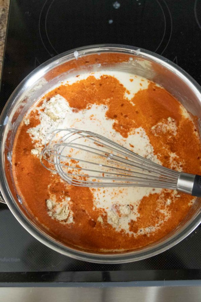 Whisk mixing red spices, cream, and thickening agent in a silver pan on a stovetop.