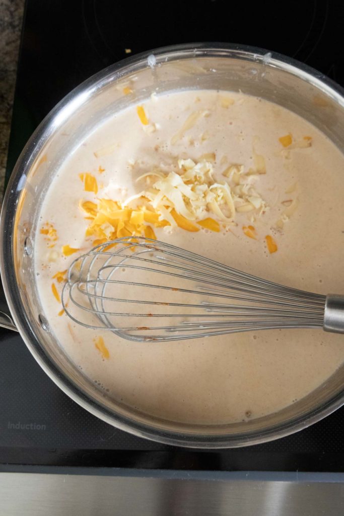 A saucepan on a stovetop contains a creamy mixture with shredded cheese and a whisk.