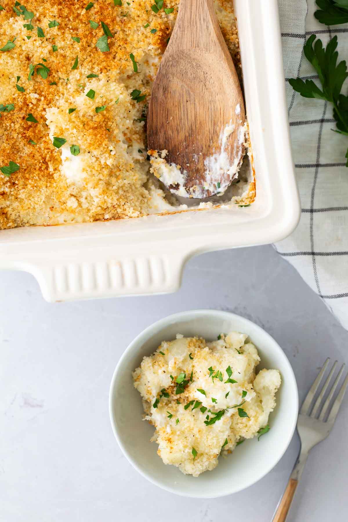 Baked dish with golden breadcrumbs in a ceramic dish; a wooden spoon serves a portion into a bowl. Garnished with parsley, a fork rests nearby on a checkered cloth.