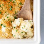 A white baking dish with a wooden spoon scooping into a cauliflower and breadcrumb casserole, garnished with chopped herbs.