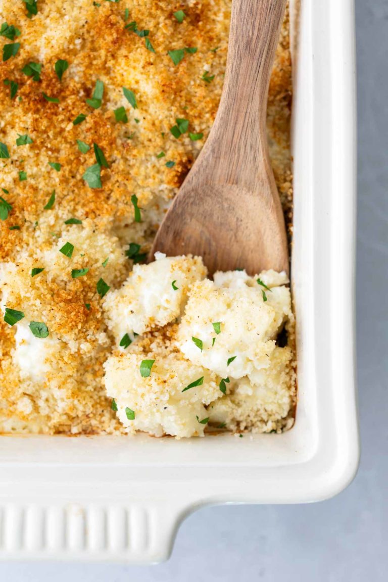 A white baking dish with a wooden spoon scooping into a cauliflower and breadcrumb casserole, garnished with chopped herbs.