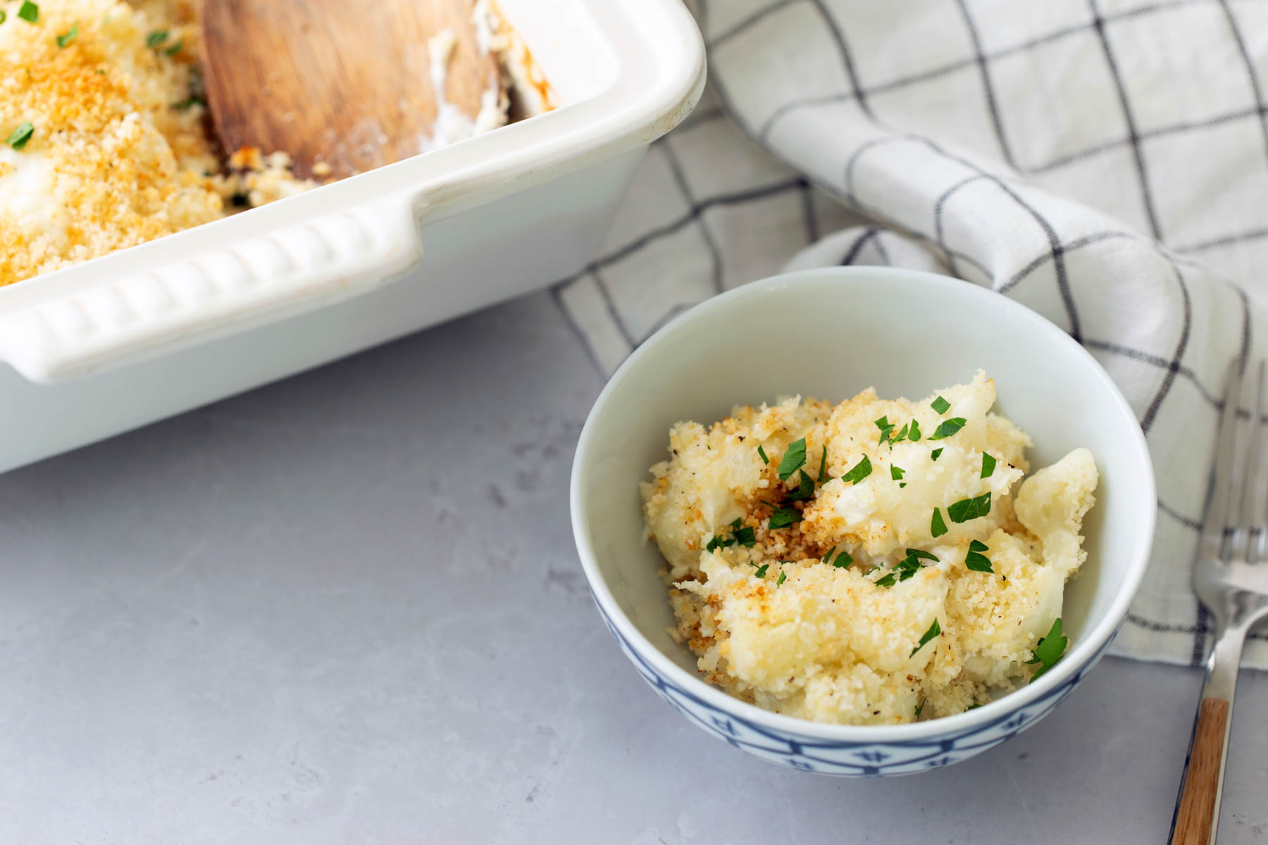 A bowl of baked cauliflower with breadcrumbs and herbs beside a partially served casserole dish on a gray surface with a checkered cloth.