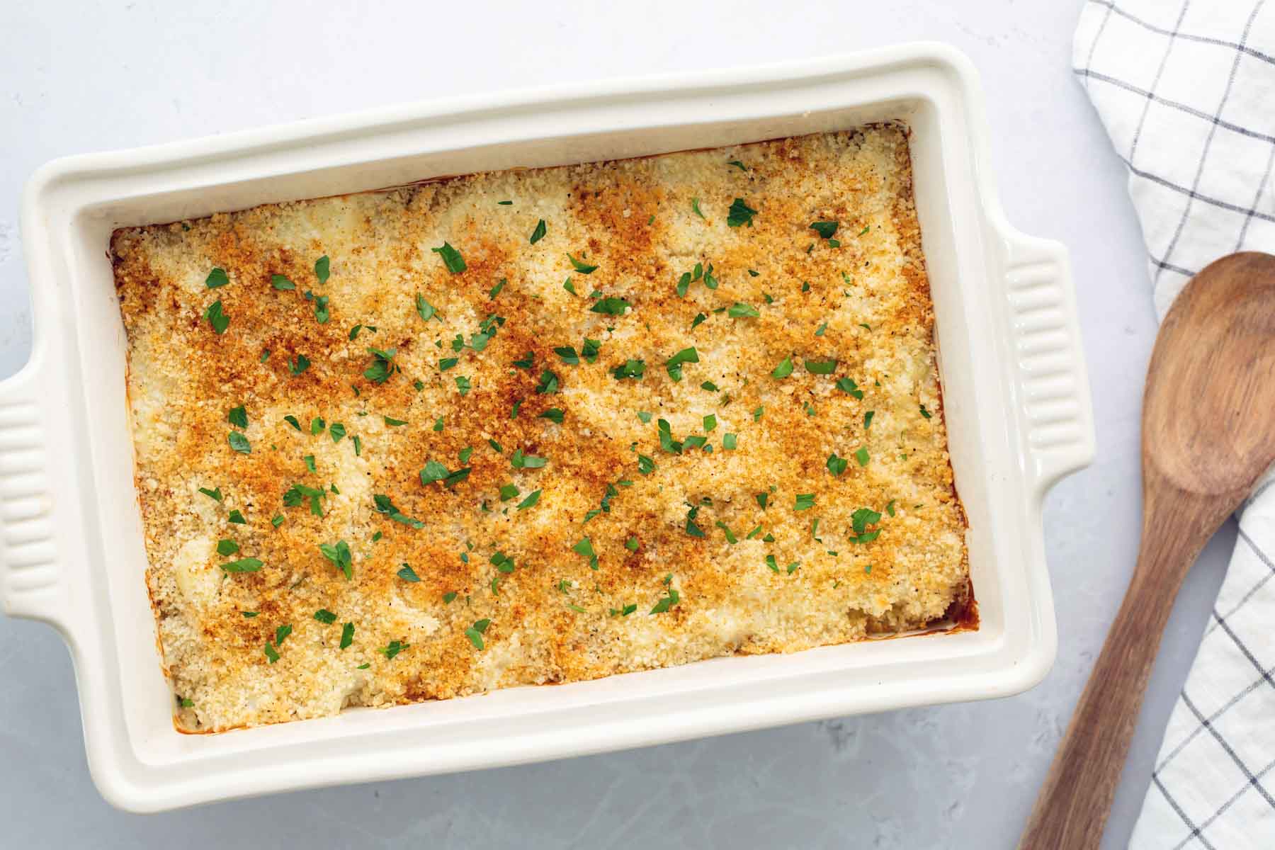 A casserole dish with a baked crispy topping, garnished with herbs, sits next to a wooden spoon and checkered cloth.