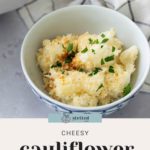 A bowl of cheesy cauliflower gratin topped with breadcrumbs and parsley, placed on a light gray surface with a checkered cloth nearby.