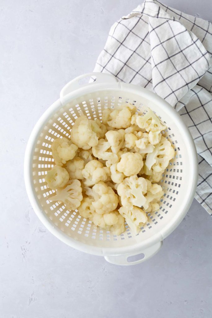 A white colander filled with cauliflower florets next to a checkered dish towel on a light surface.