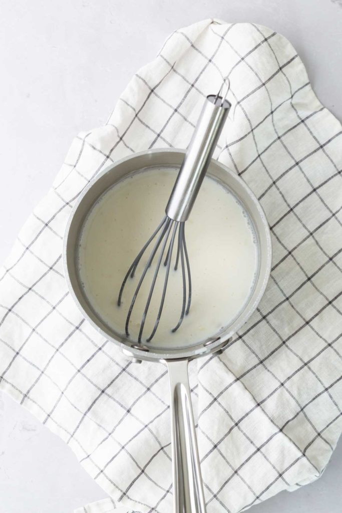 A saucepan with white sauce and a whisk inside is placed on a checkered cloth.