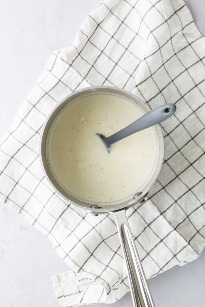 A saucepan with creamy white sauce and a stirring spoon inside, placed on a checkered cloth.