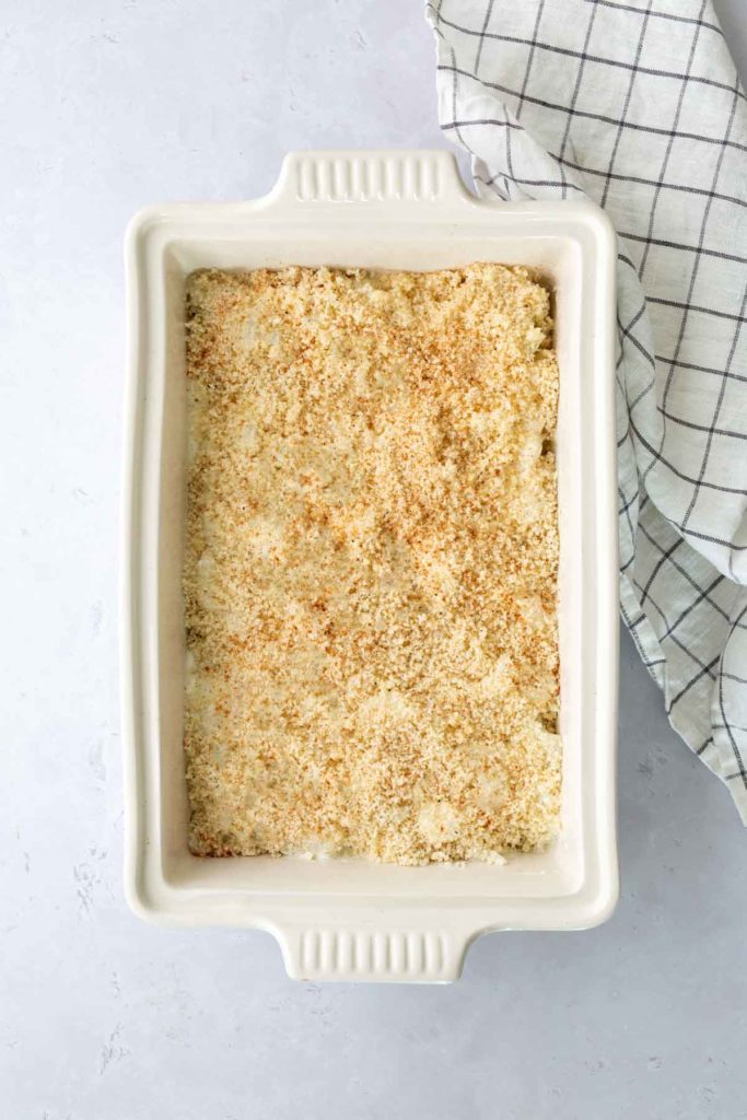 A rectangular baking dish filled with breadcrumb-topped casserole sits next to a folded checkered cloth.