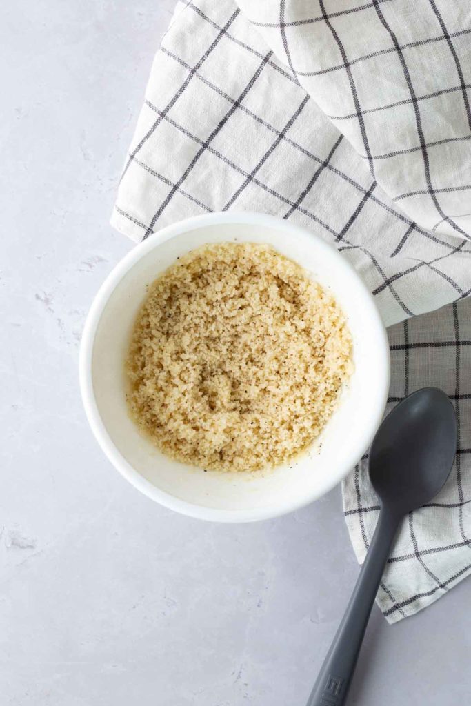 A bowl of breadcrumbs on a light surface, next to a black spoon and a checkered cloth.