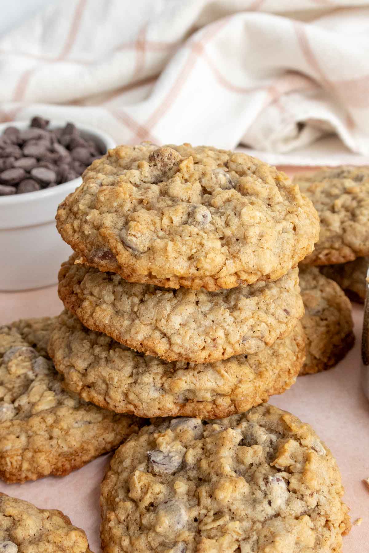 A stack of oatmeal chocolate chip cookies with a bowl of chocolate chips in the background, placed on a light-colored surface.