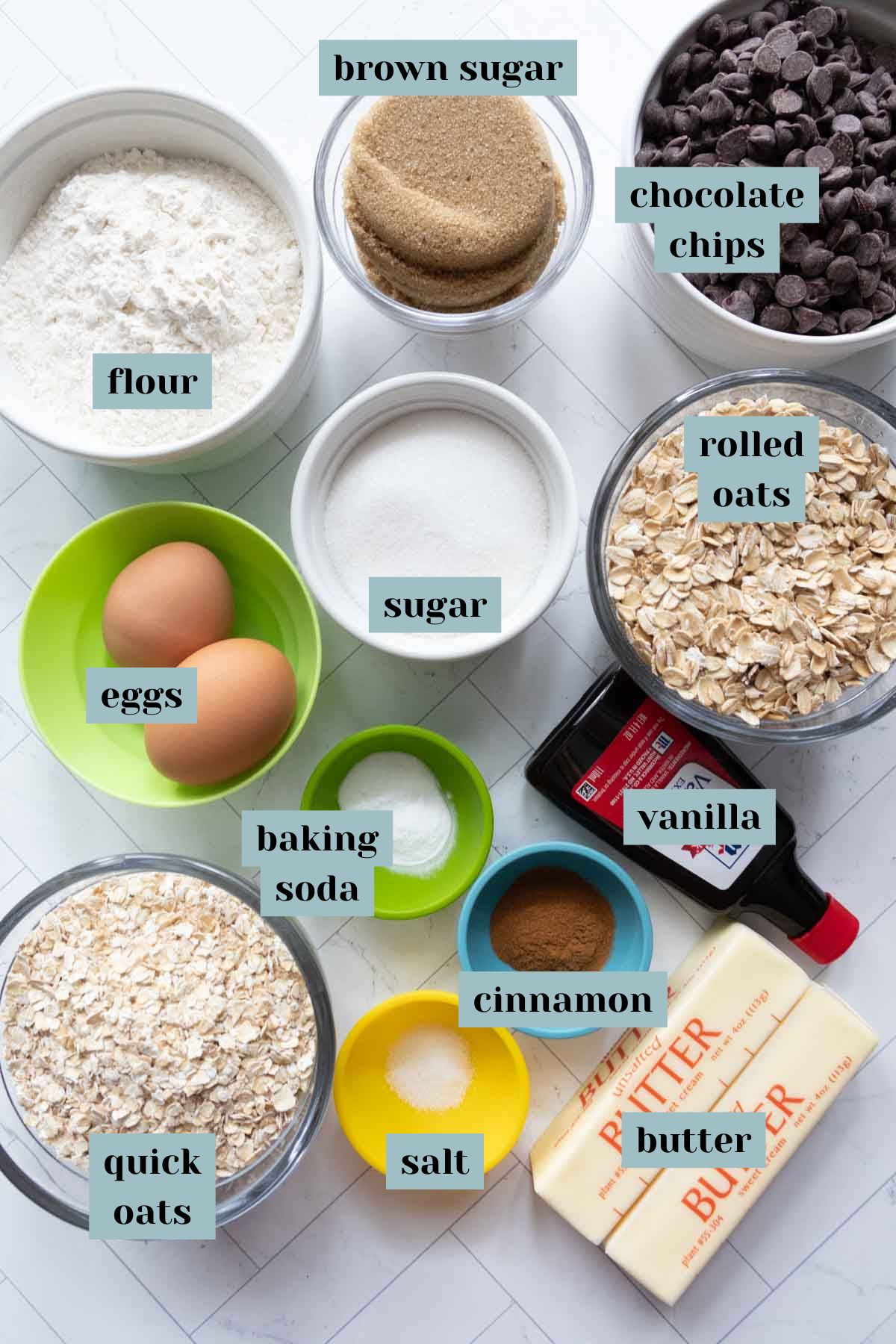 Ingredients for baking: flour, brown sugar, chocolate chips, rolled oats, quick oats, eggs, baking soda, vanilla, cinnamon, salt, butter, and regular sugar arranged on a countertop.