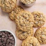 Oatmeal chocolate chip cookies are arranged on a surface, with bowls of chocolate chips and oats nearby.