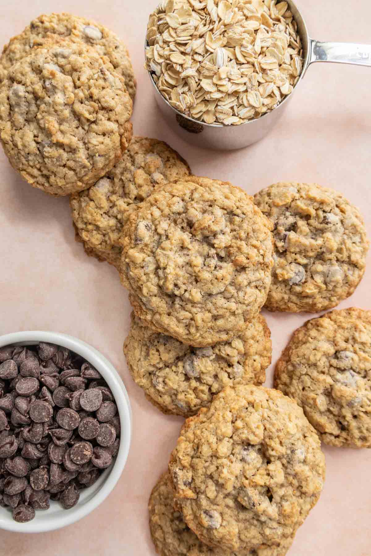 Oatmeal chocolate chip cookies are arranged on a surface, with bowls of chocolate chips and oats nearby.