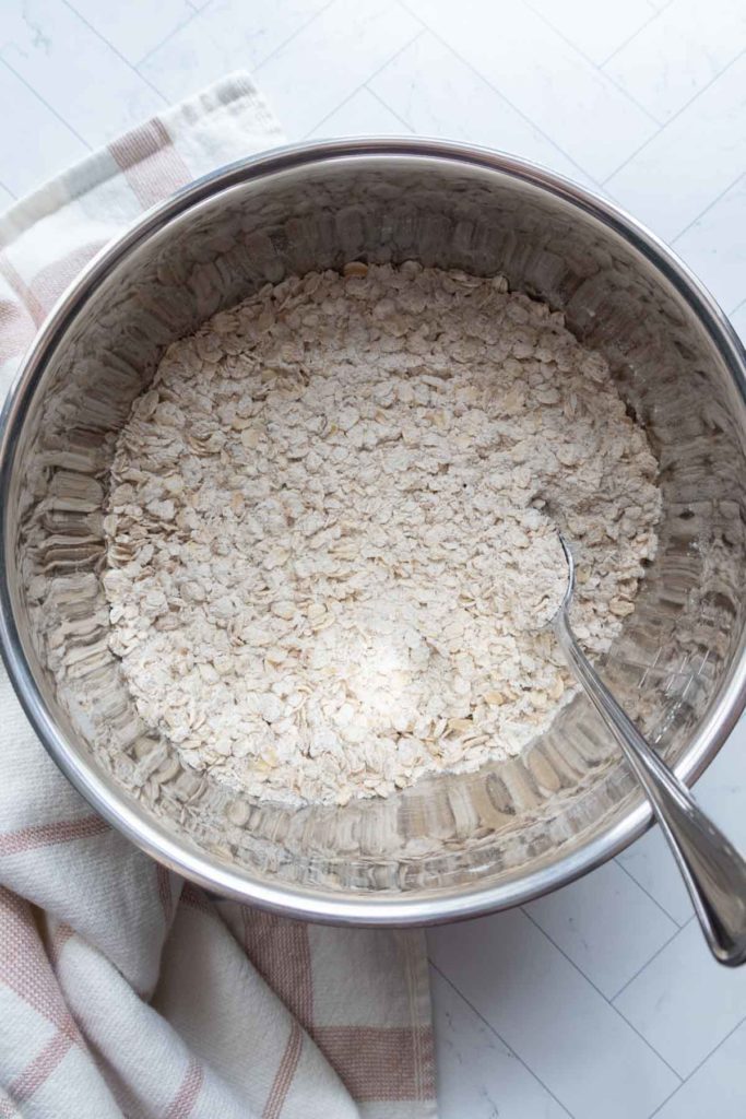 A stainless steel bowl filled with oats and flour, with a metal spoon inside. A striped cloth is partially visible beside the bowl.