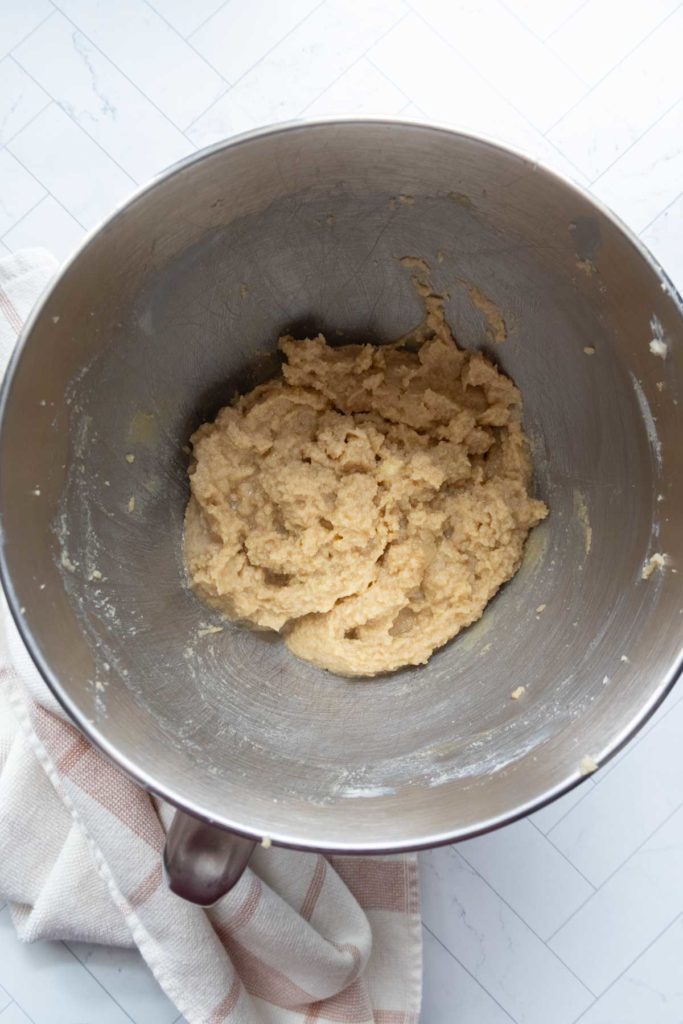 A metal mixing bowl contains raw cookie dough on a white surface with a striped cloth nearby.