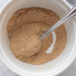 A white bowl filled with brownish powder and a metal spoon resting inside, placed on a hexagonal patterned surface.