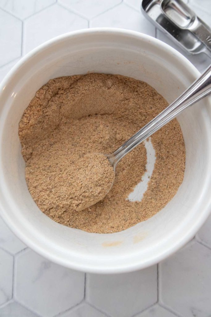 A white bowl filled with brownish powder and a metal spoon resting inside, placed on a hexagonal patterned surface.