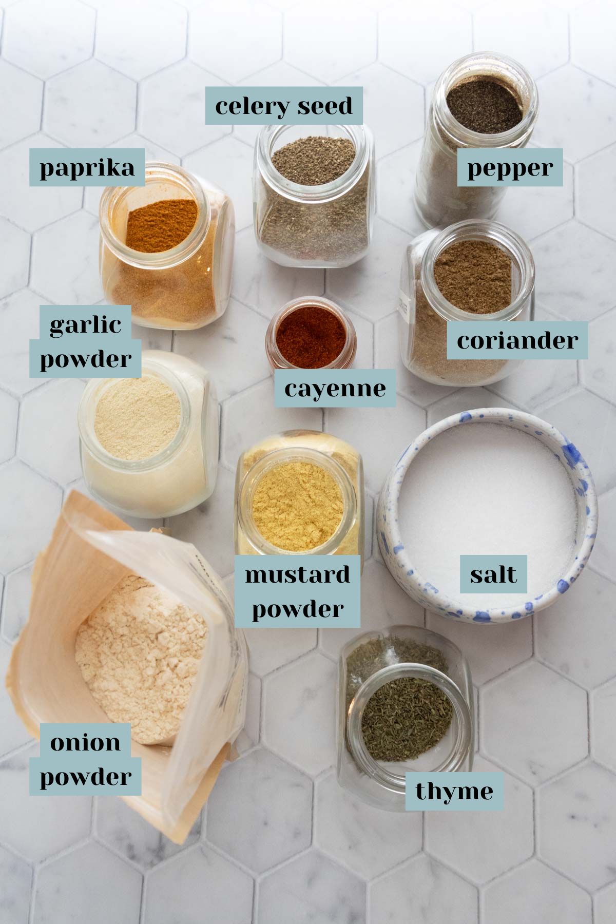 Various spices and herbs in jars labeled as paprika, pepper, garlic powder, coriander, cayenne, celery seed, onion powder, mustard powder, salt, and thyme on a white hexagonal tiled surface.