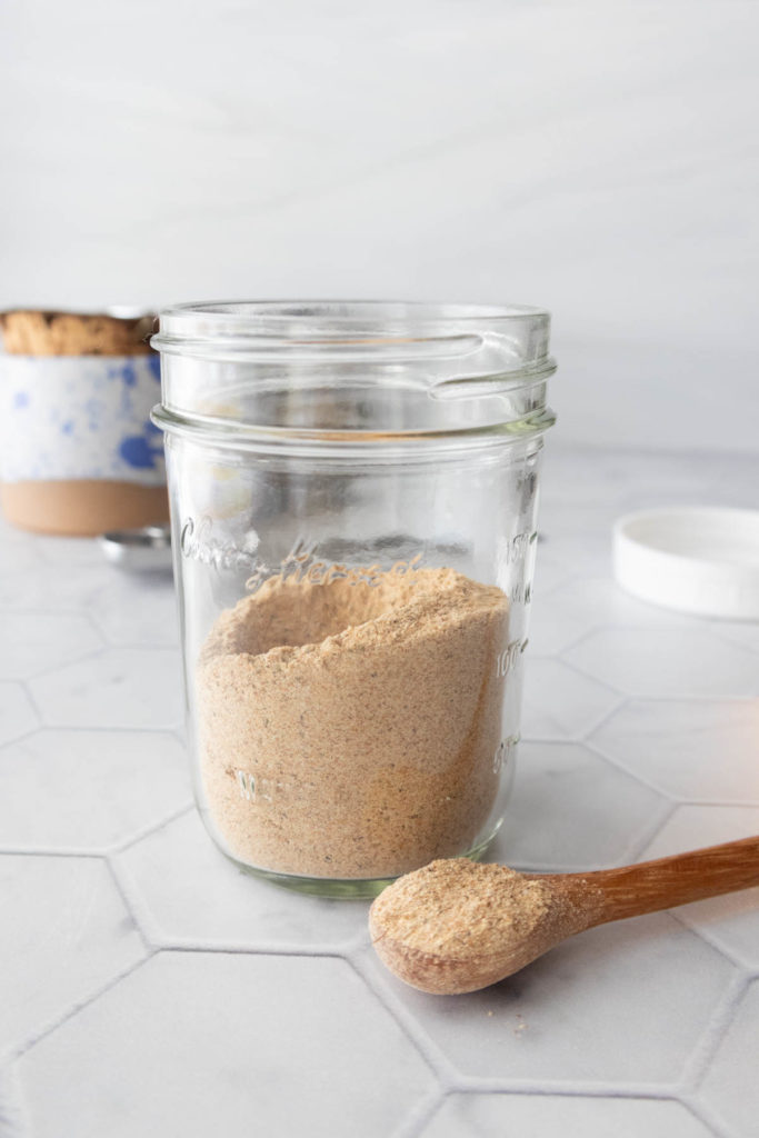 A glass jar filled with a light brown powder, next to a wooden spoon holding some powder, on a hexagonal-tiled surface. A blurred cup is in the background.