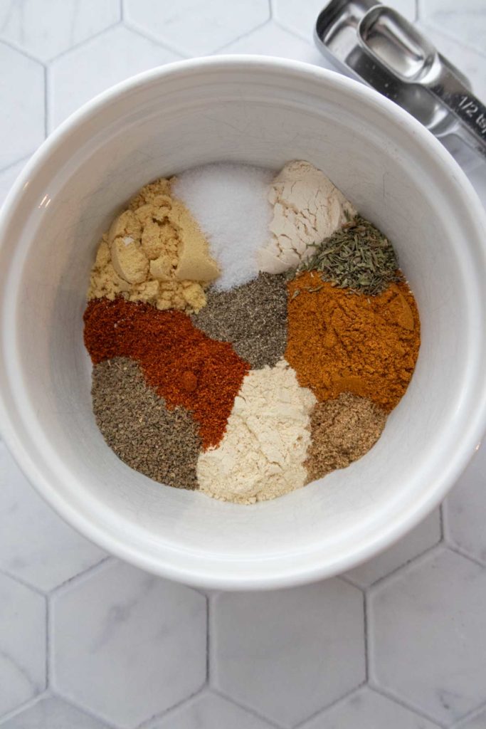 Overhead view of a bowl containing various spices in sections, placed on a hexagonal tile surface. Measuring spoons are visible beside the bowl.