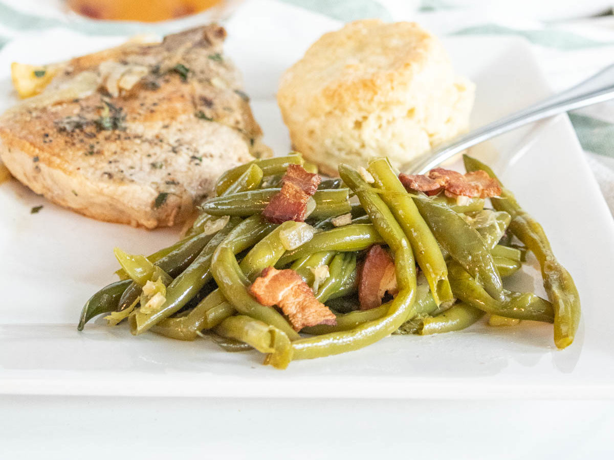 Dinner plate with southern green beans alongside a pork chop and biscuit with a glass of sangria in the background.