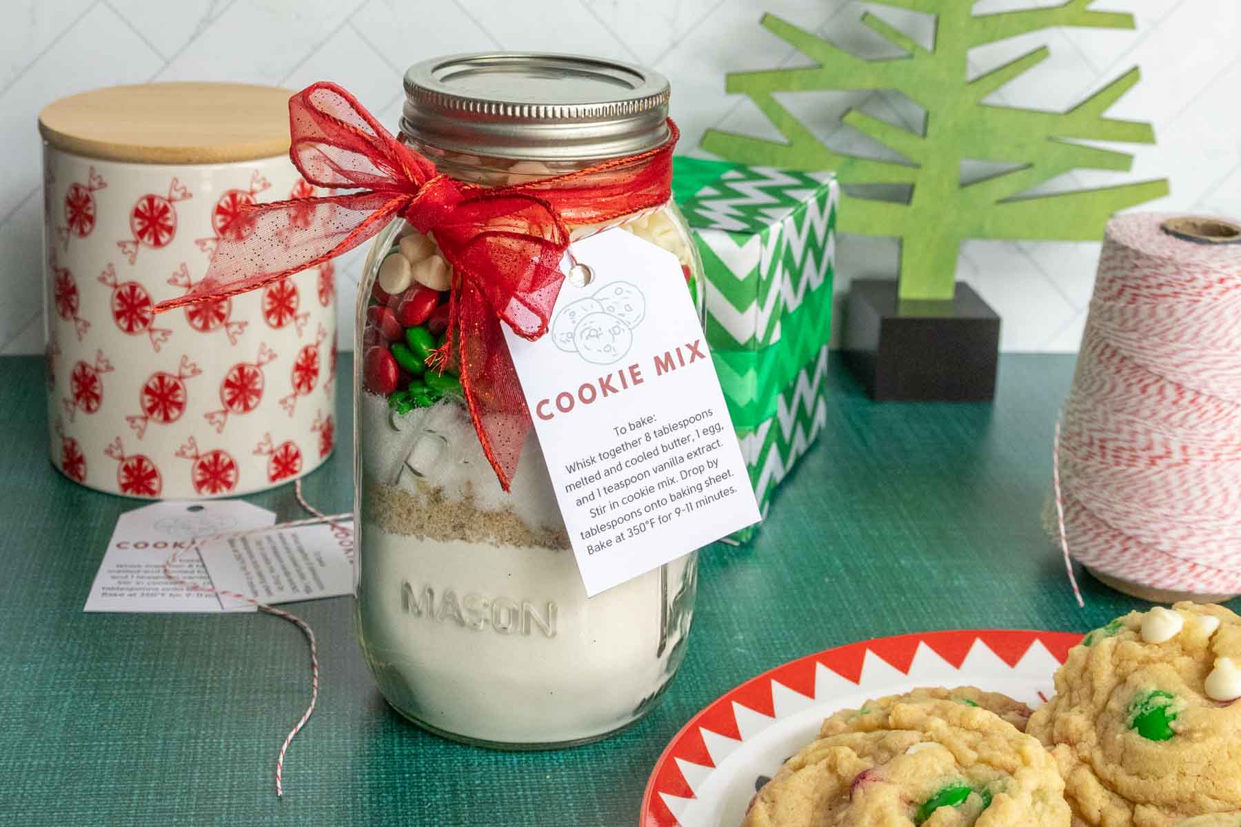 A jar of cookie mix with a red ribbon and tag sits next to a plate of baked cookies and decorative items on a green surface.