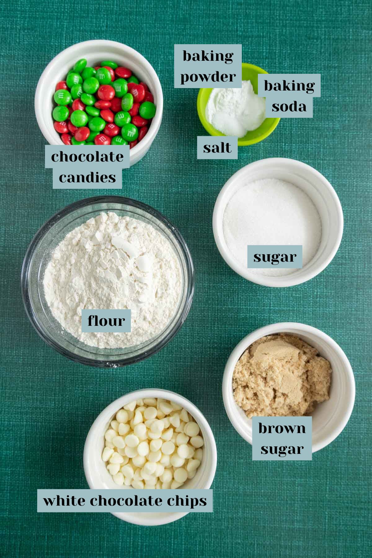 Ingredients for a cookie recipe laid out on a green surface, including chocolate candies, baking powder, baking soda, salt, sugar, flour, brown sugar, and white chocolate chips.