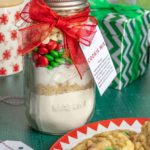 A mason jar filled with cookie mix, topped with red and green candies, and tied with a red ribbon and tag. In the background are gifts, and a plate of cookies sits in the foreground.