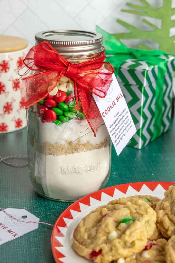 A mason jar filled with cookie mix, topped with red and green candies, and tied with a red ribbon and tag. In the background are gifts, and a plate of cookies sits in the foreground.