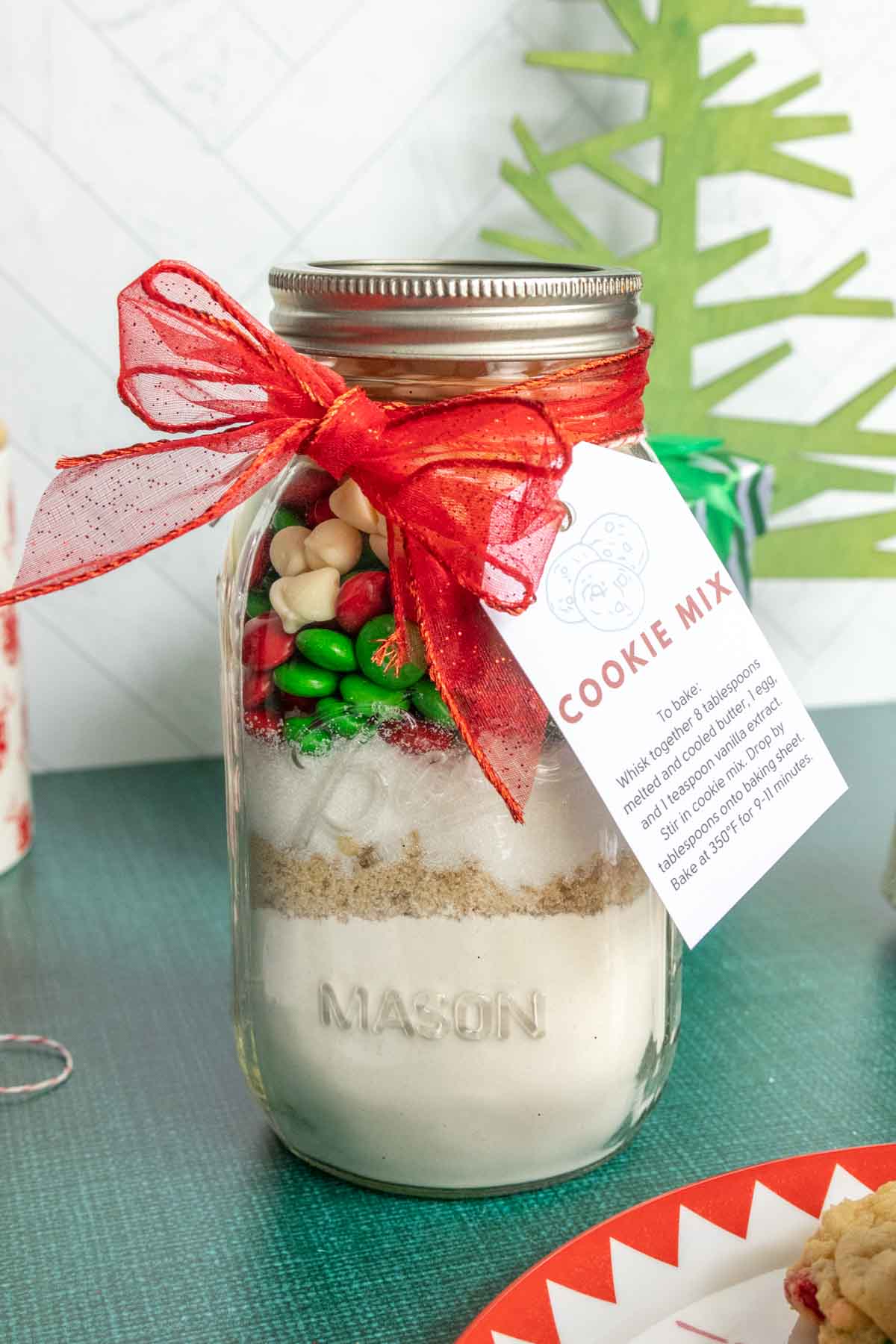 A mason jar filled with layered cookie mix ingredients, topped with festive candies, and tied with a red ribbon. A tag with baking instructions is attached.