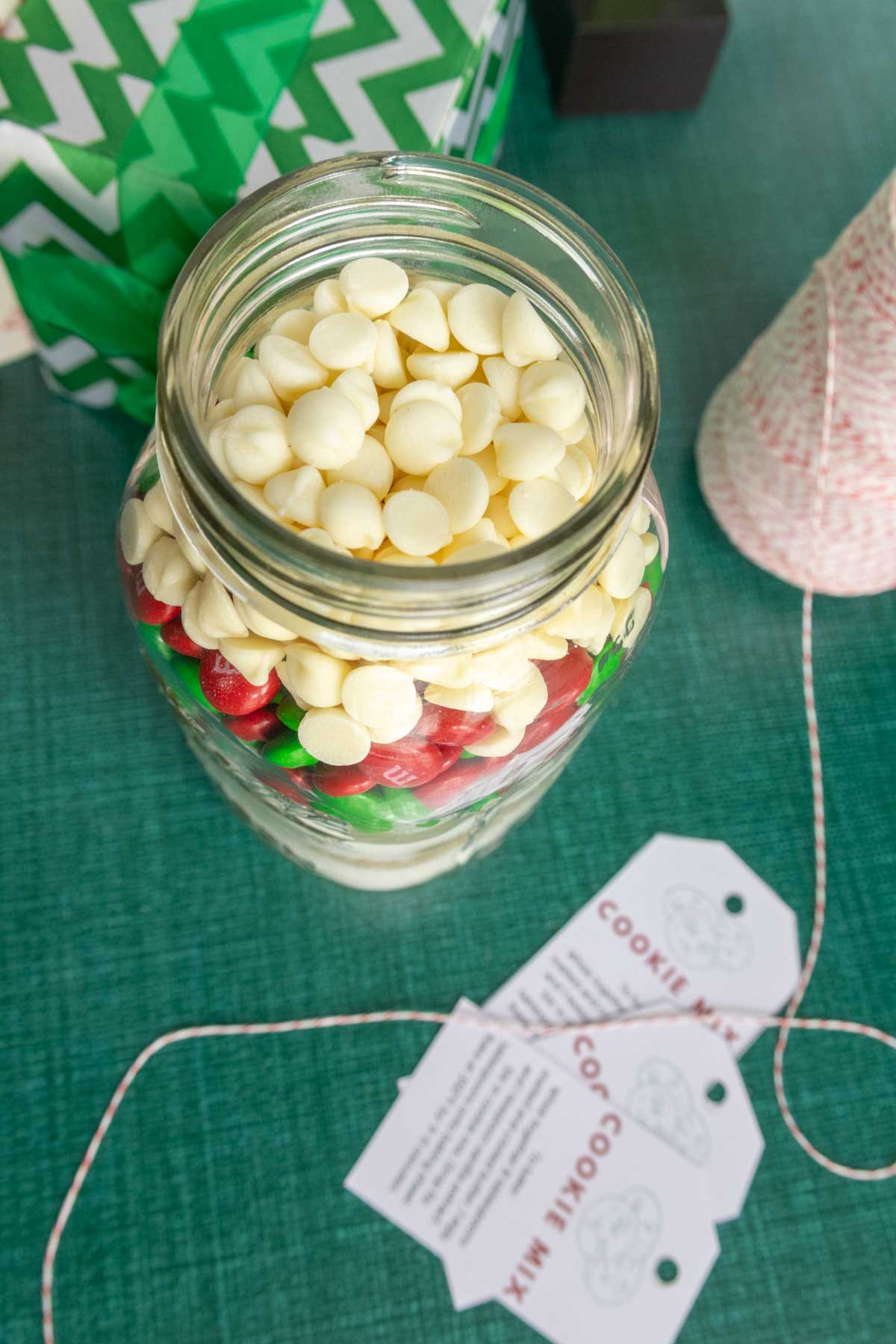 A jar filled with layers of white chocolate chips, red, and green candies on a green table, next to a roll of red and white string and three cookie mix tags.