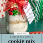 Mason jar with layered cookie mix ingredients, topped with a red ribbon. A gift label is attached. Decorated Christmas gifts are in the background.