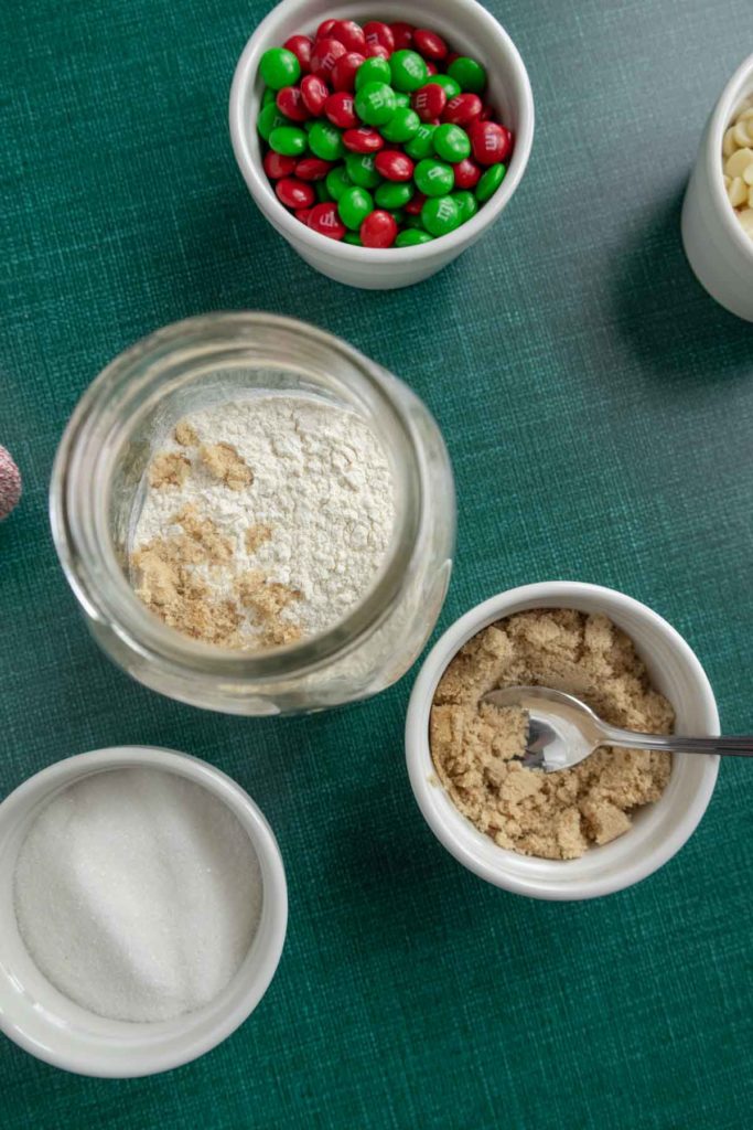 Ingredients for baking on a green surface, including a jar with flour and brown sugar, a small bowl of sugar, another bowl with brown sugar and a spoon, and a bowl of red and green candies.