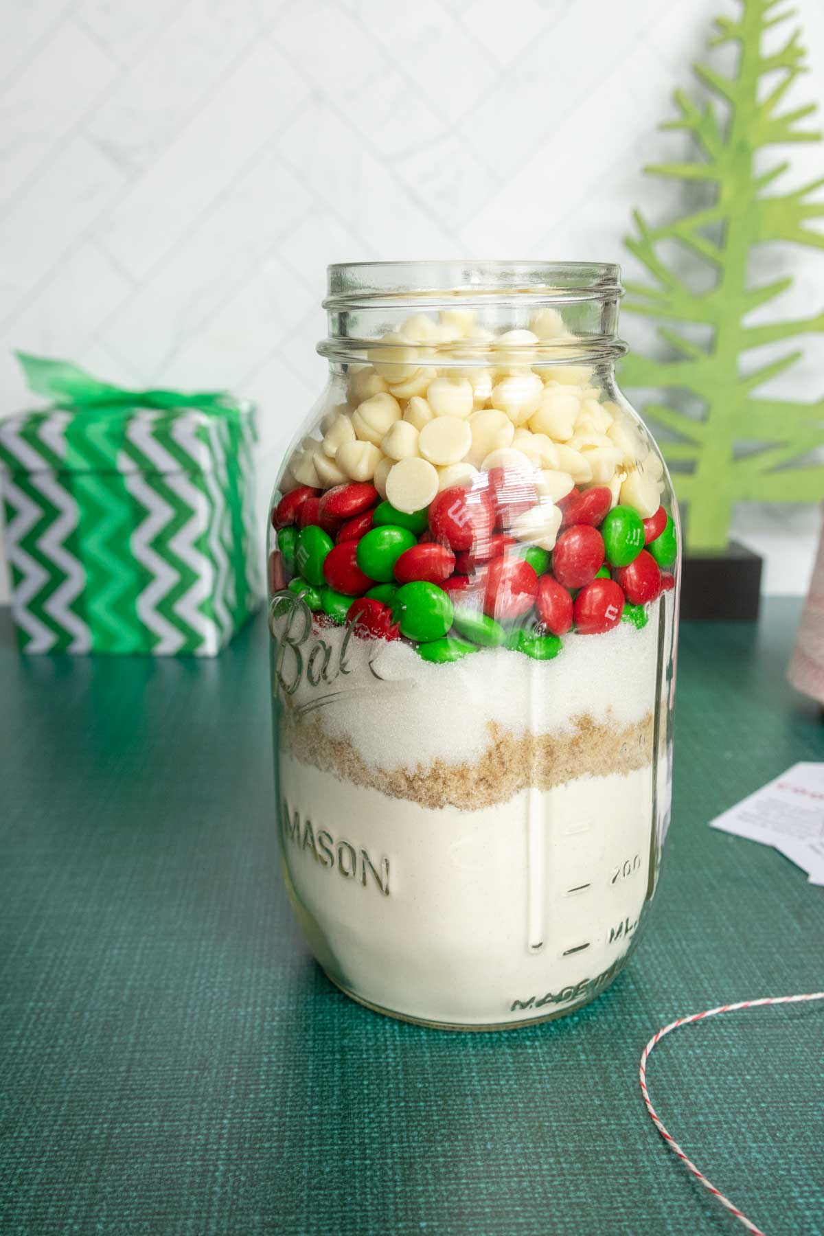 A mason jar filled with white chocolate chips, red and green candies, brown sugar, and flour. A green tree decoration and wrapped gift are in the background.