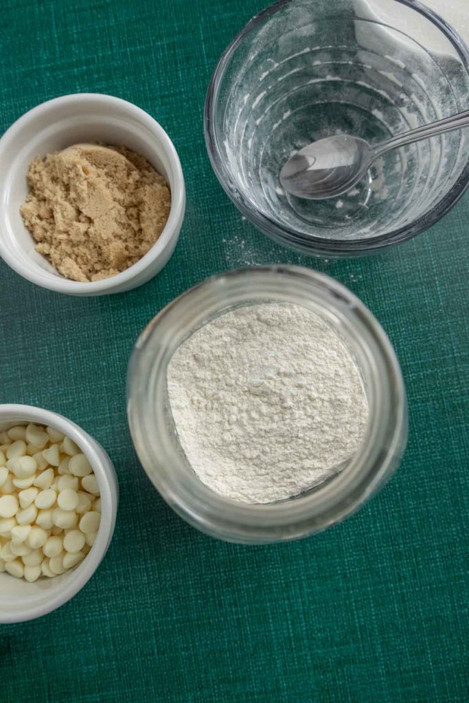 Top-down view of flour, brown sugar, white chocolate chips in small bowls, and an empty glass bowl with a metal spoon on a green surface.