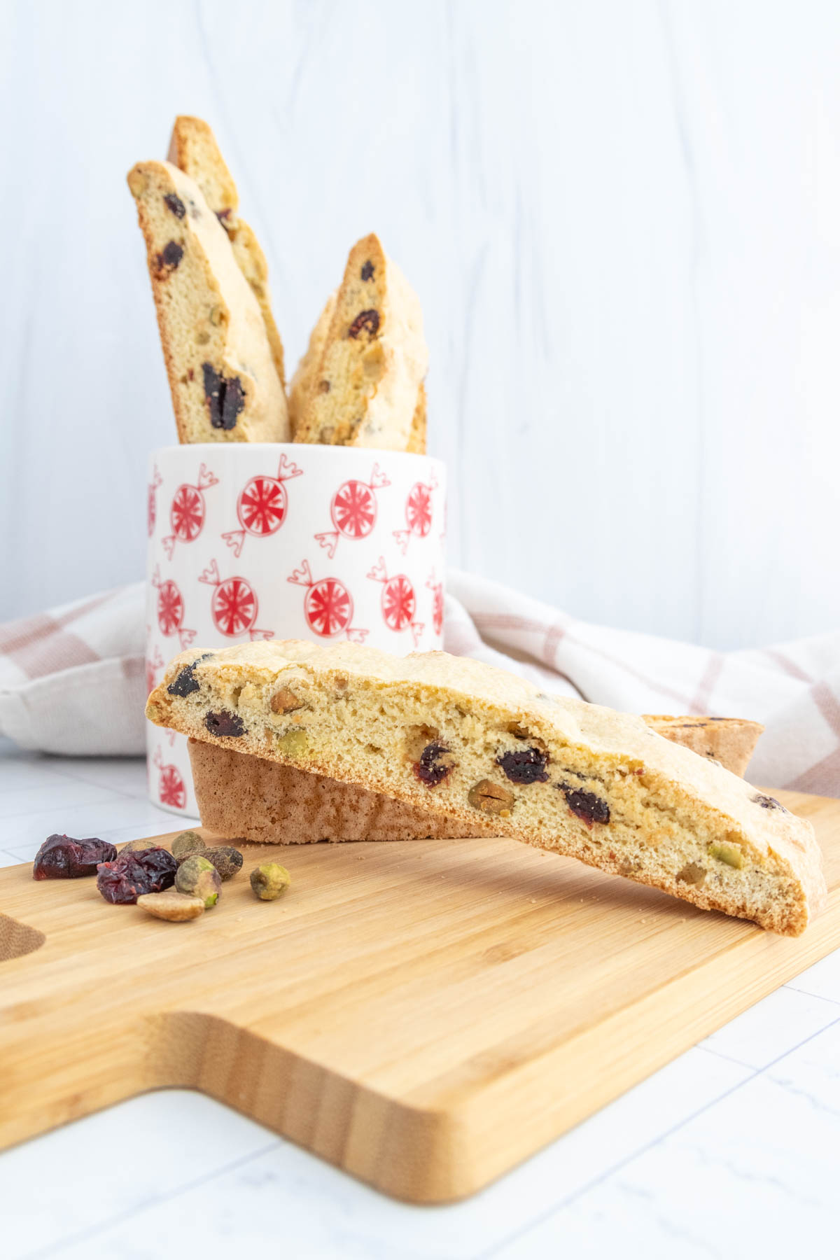 Biscotti with visible cranberries and pistachios are placed on a wooden cutting board. A patterned cup holds additional biscotti in the background.