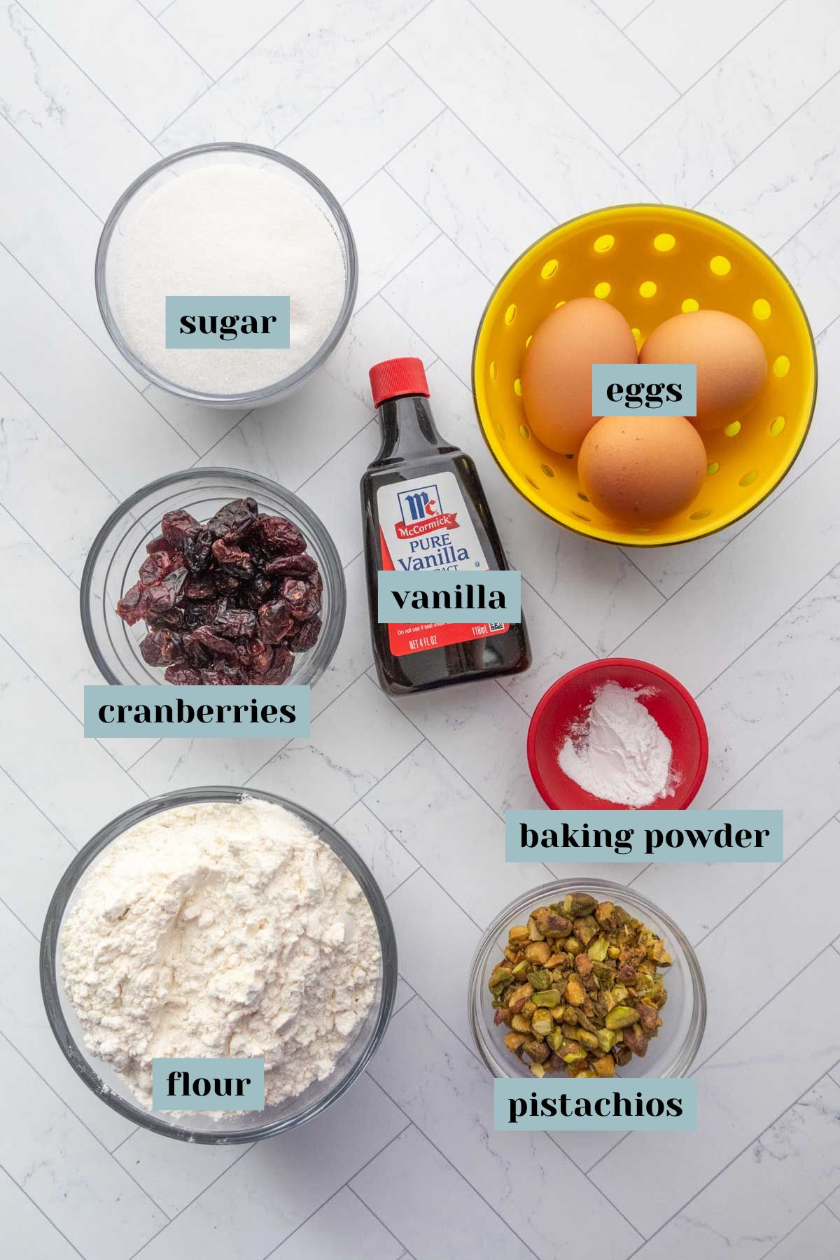 Ingredients displayed on a white surface: sugar, eggs in a yellow bowl, vanilla extract, cranberries, baking powder, flour, and pistachios, each labeled.