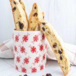 Biscotti with nuts and dried fruits in a decorative mug, surrounded by scattered nuts and dried fruits on a white surface.