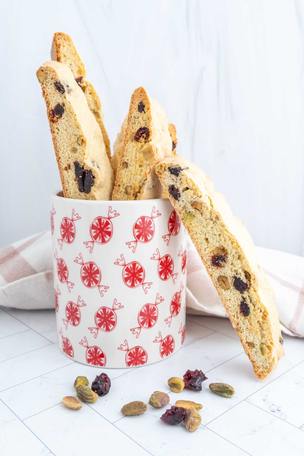 Biscotti with nuts and dried fruits in a decorative mug, surrounded by scattered nuts and dried fruits on a white surface.